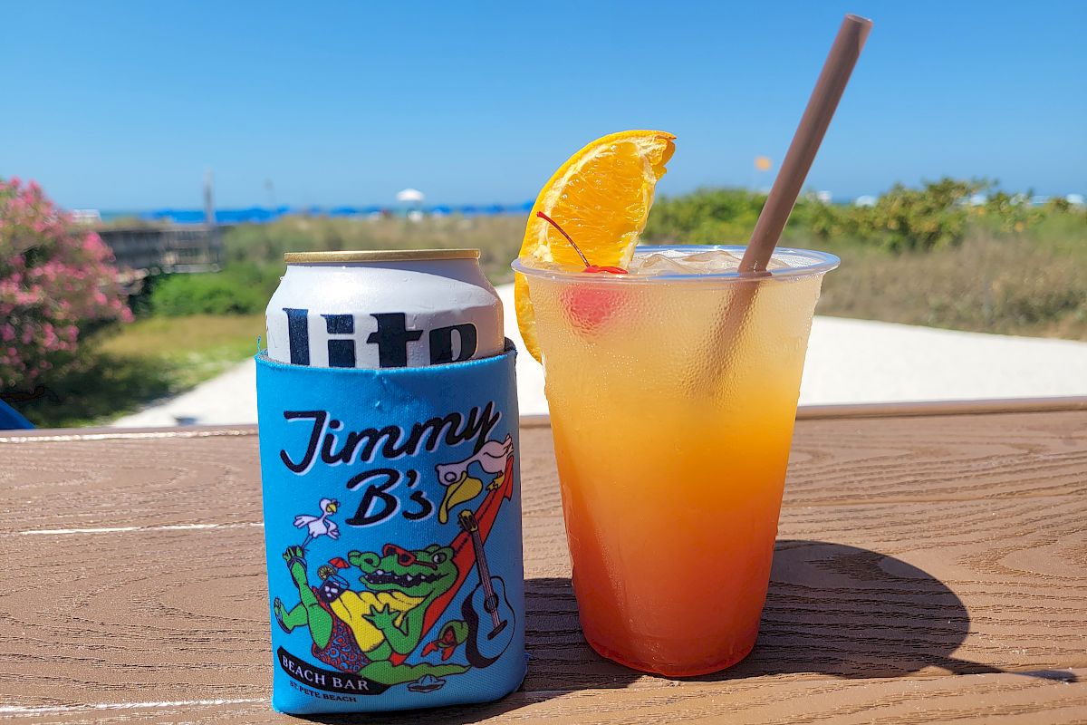A colorful can of &ldquo;Ittro Jimmy B&rdquo; and a tropical orange-pink drink with a lime/orange wedge on a wooden table, sunny outdoor scene.