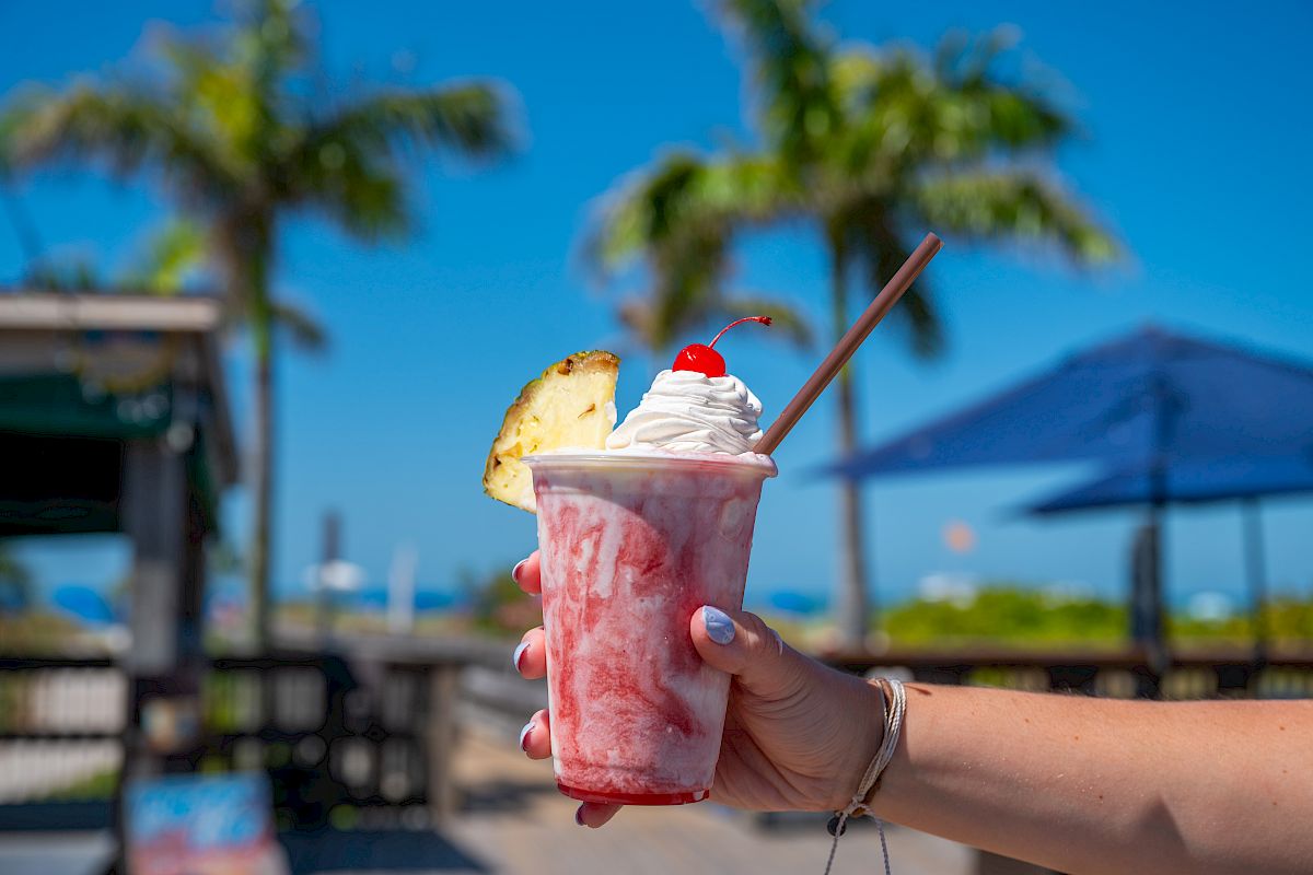 A hand holding a pink berry smoothie drink topped with whipped cream, a cherry, and a pineapple slice, outdoors on a sunny day with palm trees.