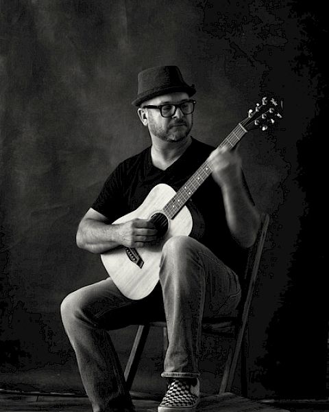 A person wearing a hat and glasses, sitting on a chair, playing an acoustic guitar, with one foot resting on a wooden block, in a black and white photo.