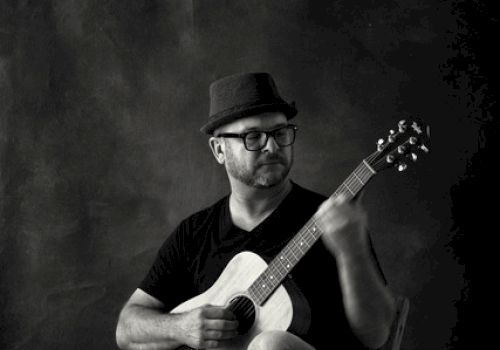 A person wearing a hat and glasses, sitting on a chair, playing an acoustic guitar, with one foot resting on a wooden block, in a black and white photo.