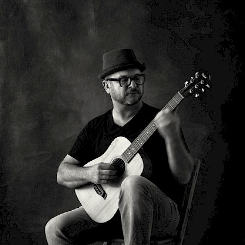 A person wearing a hat and glasses, sitting on a chair, playing an acoustic guitar, with one foot resting on a wooden block, in a black and white photo.