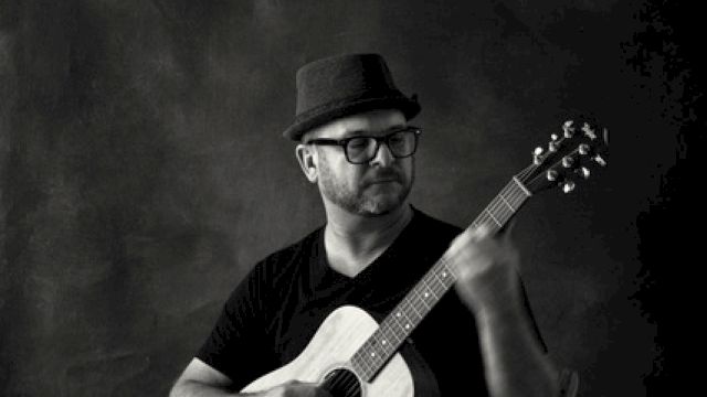 A person wearing a hat and glasses, sitting on a chair, playing an acoustic guitar, with one foot resting on a wooden block, in a black and white photo.