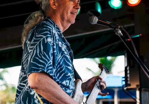 A person with long hair performs on stage, playing a guitar and standing in front of a microphone, under colorful stage lights.