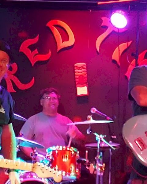 A band performing on stage with two guitarists and a drummer, all smiling and lit by stage lights.