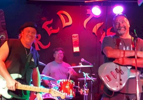 A band performing on stage with two guitarists and a drummer, all smiling and lit by stage lights.