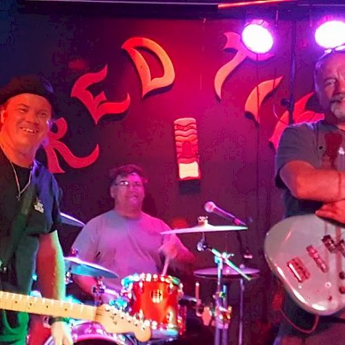 A band performing on stage with two guitarists and a drummer, all smiling and lit by stage lights.