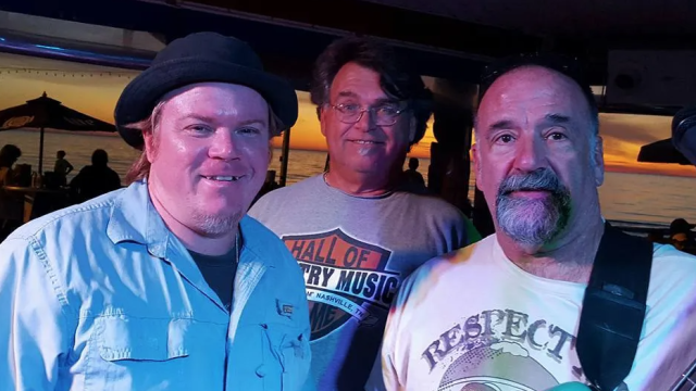 Three men are posing inside a venue, with sunset views outside. They are dressed casually and one is holding a guitar.
