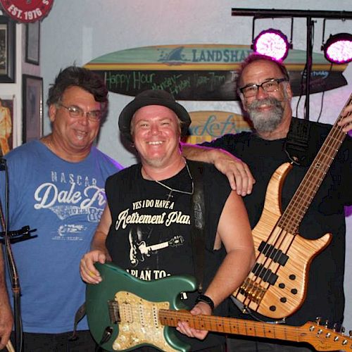 Three men are posing happily; two of them are holding musical instruments (a guitar and a bass) in a room resembling a music studio.