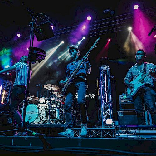 A band performing on stage with colorful lighting, featuring guitarists, a bassist, and a drummer, creating a lively and energetic atmosphere.