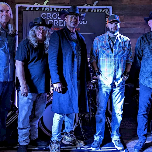 Five men are standing in a line on a stage with instruments behind them, under blue lighting, dressed in casual and cowboy attire.