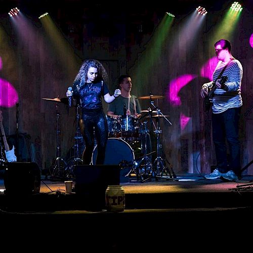 A band is performing on stage with colorful lighting; it includes a singer, two guitarists, a bassist, and a drummer, the audience in front.