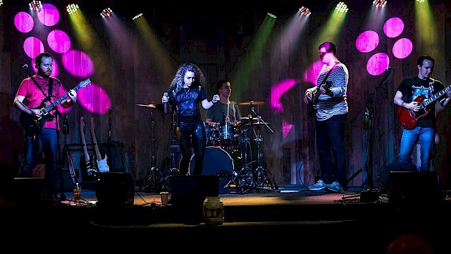 A band is performing on stage with colorful lighting; it includes a singer, two guitarists, a bassist, and a drummer, the audience in front.