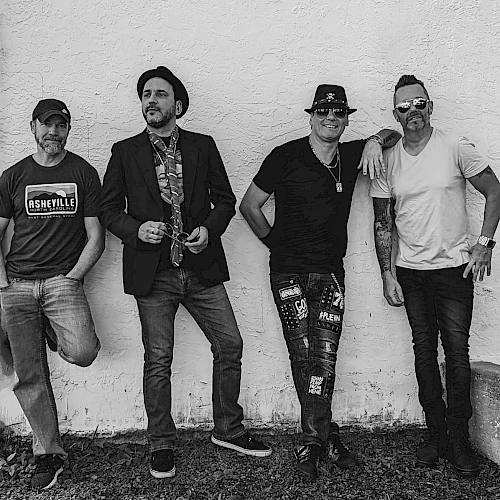 A black and white photo of four men standing against a wall. They are casually dressed and appear relaxed, with two wearing hats.