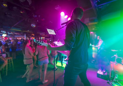 A musician performs with a guitar on stage under vibrant lighting as the audience enjoys the show at a lively, crowded venue.