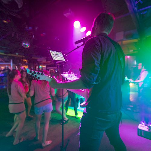 A musician performs with a guitar on stage under vibrant lighting as the audience enjoys the show at a lively, crowded venue.