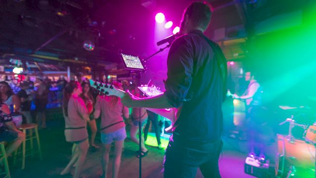A musician performs with a guitar on stage under vibrant lighting as the audience enjoys the show at a lively, crowded venue.