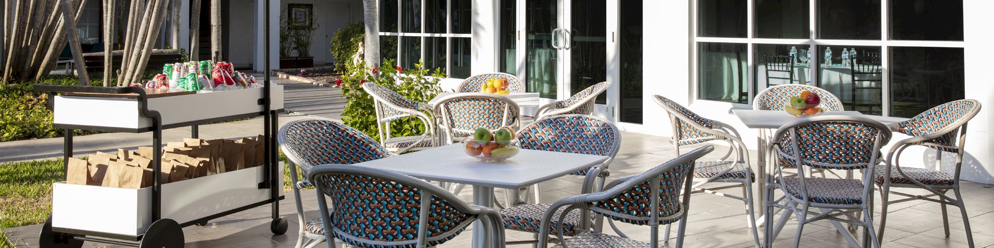 An outdoor patio with tables and chairs, a black-and-white striped umbrella, and a cart with crates, set against a white building with large windows.