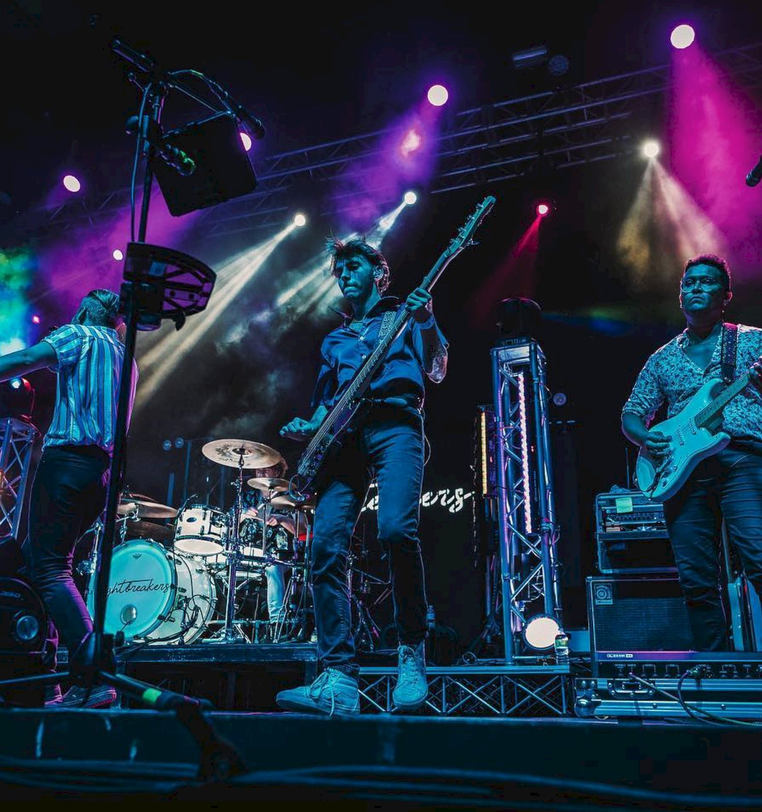 A band performs on stage with vibrant, colorful lights in the background. The members play various instruments, including guitars and drums.