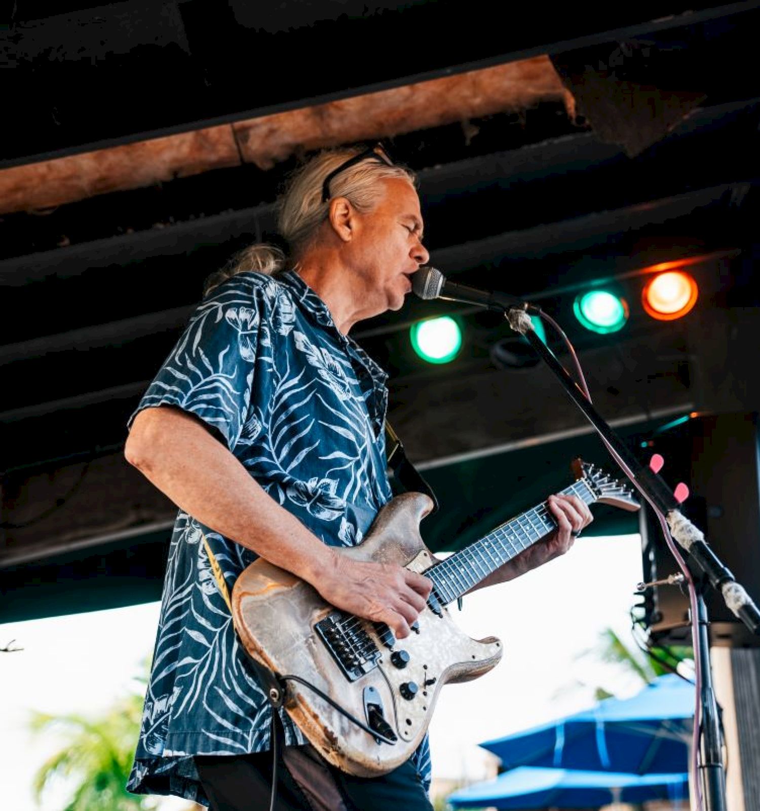 A guitarist is performing on an outdoor stage, wearing a blue patterned shirt, with colorful stage lights above him and a tropical background.