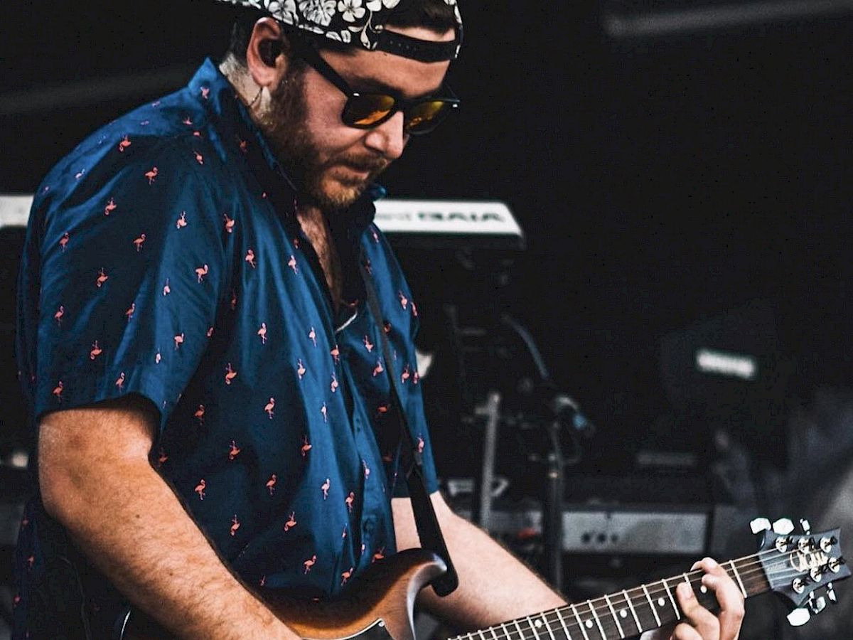 A man wearing a blue shirt and sunglasses plays an electric guitar on stage, with musical equipment visible in the background.