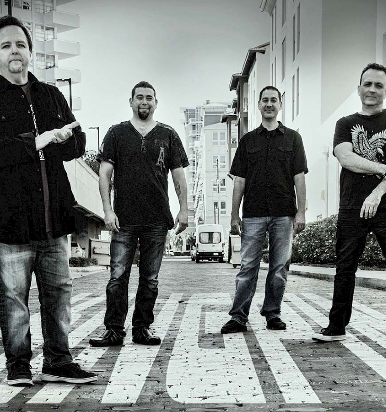 Four men stand on a street with buildings in the background, posing for the camera in a black-and-white photo.