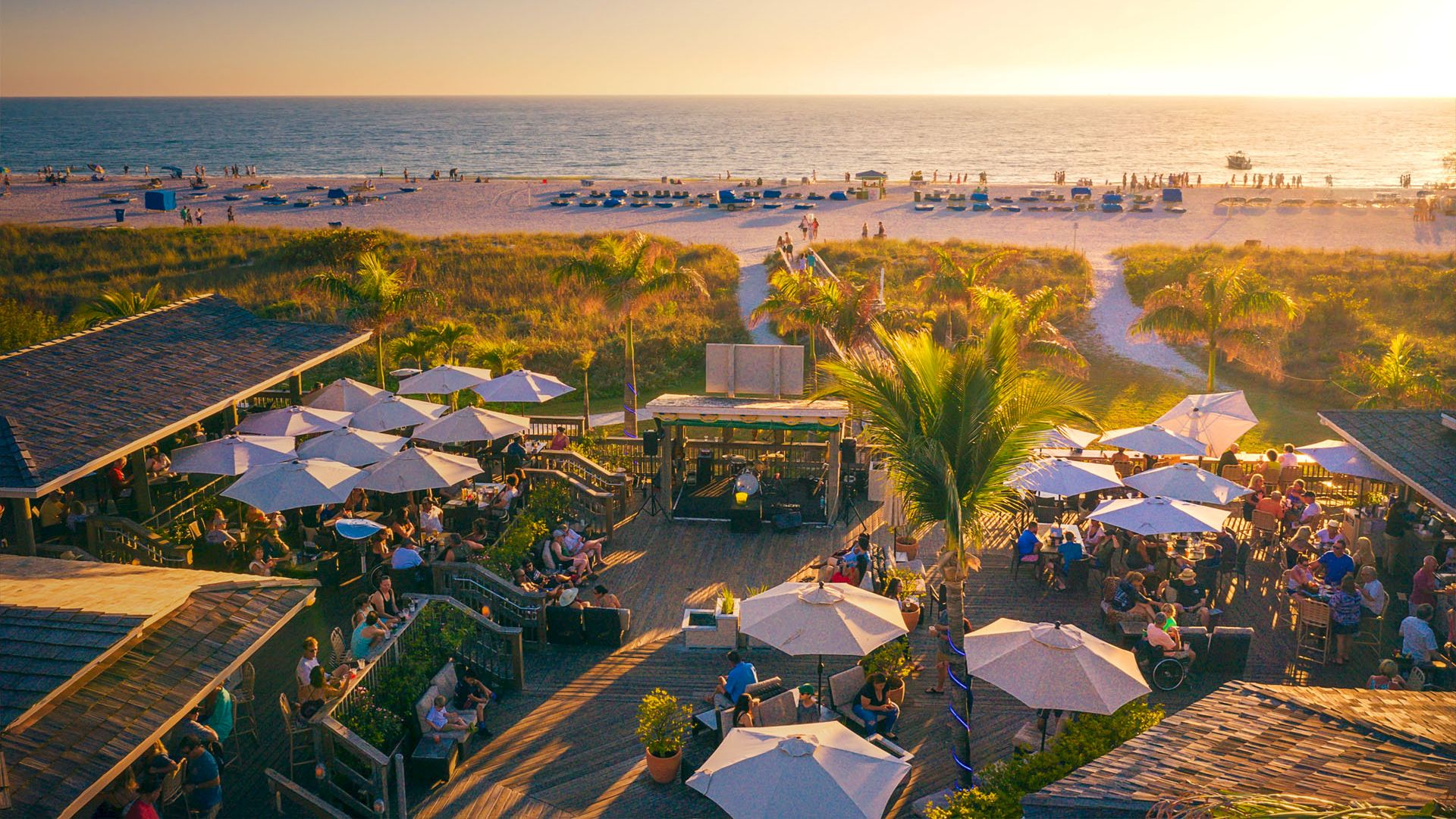 A beachside scene with people enjoying food and drinks under umbrellas, with the sandy shore and ocean in the background at sunset.
