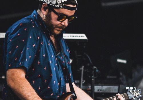 A person wearing sunglasses and a cap is playing an electric guitar on stage, dressed in a patterned shirt with equipment visible in the background.