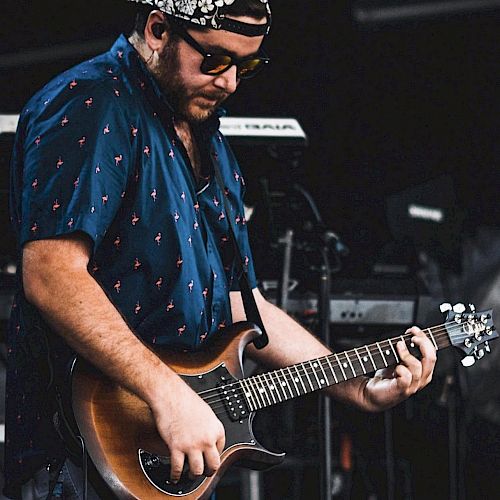 A person wearing sunglasses and a cap is playing an electric guitar on stage, dressed in a patterned shirt with equipment visible in the background.