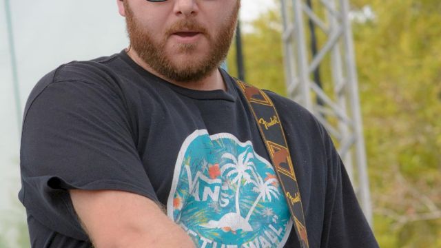 A musician is playing a guitar on stage, wearing sunglasses and a cap, with logos for "Reggae Rise Up" and "CNL" visible in the corner.