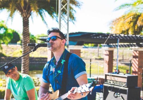 A musician is playing an acoustic guitar and singing into a microphone, with another person playing a percussion instrument, outdoors, under sunny weather.