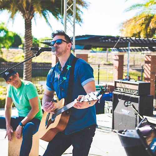 A musician is playing an acoustic guitar and singing into a microphone, with another person playing a percussion instrument, outdoors, under sunny weather.