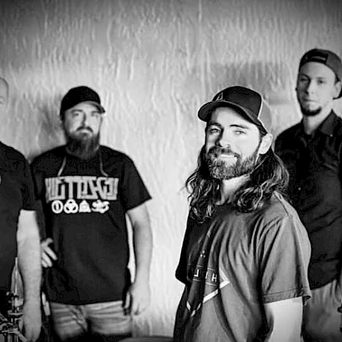 The image shows four men standing together in a black and white setting, wearing casual clothing and hats.