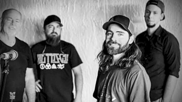 The image shows four men standing together in a black and white setting, wearing casual clothing and hats.