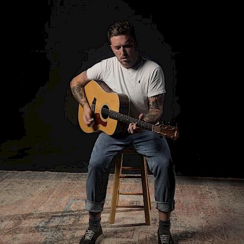 A man is sitting on a stool, playing an acoustic guitar, wearing a white shirt and jeans, with a dark background.