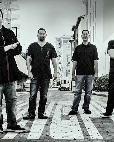 The image depicts four men standing on a city street, with buildings in the background. The photo is in black and white, and everyone faces the camera.