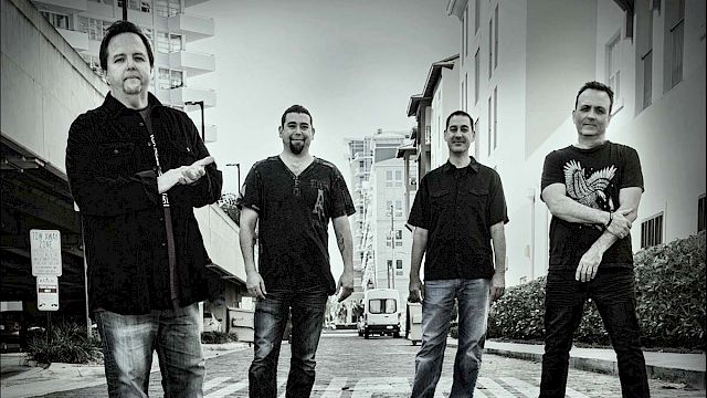 The image depicts four men standing on a city street, with buildings in the background. The photo is in black and white, and everyone faces the camera.