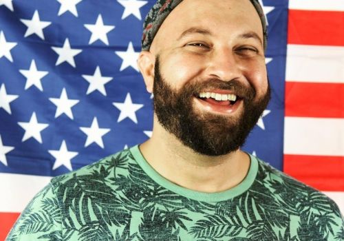 A bearded person wearing a patterned shirt and headscarf is smiling in front of an American flag backdrop.