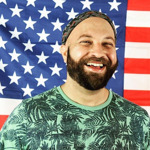 A bearded person wearing a patterned shirt and headscarf is smiling in front of an American flag backdrop.