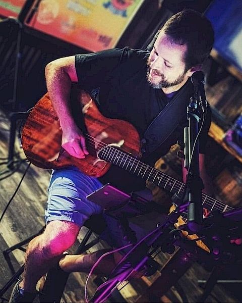 A man is playing an acoustic guitar, seated in a dimly lit room with musical equipment around them. They appear to be performing or practicing.