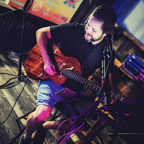 A man is playing an acoustic guitar, seated in a dimly lit room with musical equipment around them. They appear to be performing or practicing.