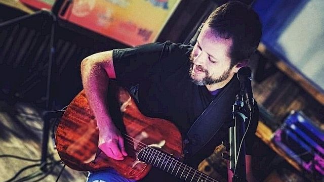 A man is playing an acoustic guitar, seated in a dimly lit room with musical equipment around them. They appear to be performing or practicing.