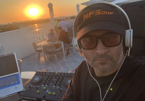 A man is wearing headphones and a cap, standing near DJ equipment, with a sunset and people sitting at tables in the background.