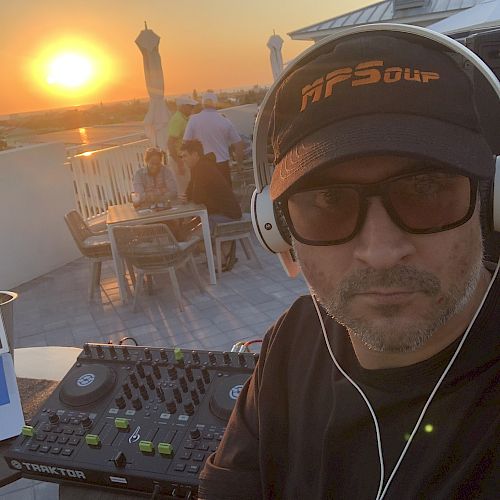 A man is wearing headphones and a cap, standing near DJ equipment, with a sunset and people sitting at tables in the background.