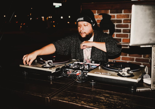 The image shows a DJ wearing a beanie and headphones, performing with turntables and a laptop, in front of a brick wall backdrop.