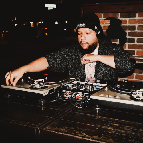 The image shows a DJ wearing a beanie and headphones, performing with turntables and a laptop, in front of a brick wall backdrop.