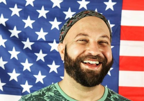 A smiling bearded man stands in front of an American flag, wearing a green shirt with a leafy pattern and a headband.