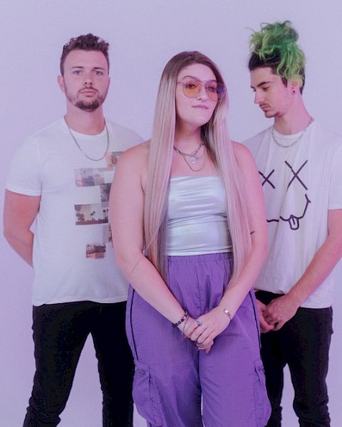 Three individuals are posing in front of a plain background. They are casually dressed, with one person having green hair, and another wearing glasses.