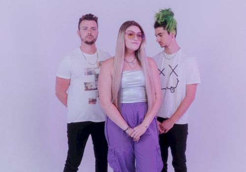 Three individuals are posing in front of a plain background. They are casually dressed, with one person having green hair, and another wearing glasses.