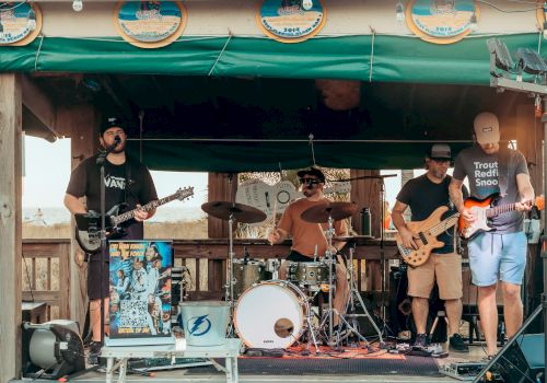 A band is performing on a small outdoor stage, with members playing guitars, drums, and bass, under a green canopy decorated with circular signs.