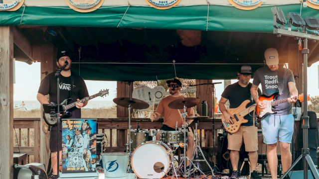 A band is performing on a small outdoor stage, with members playing guitars, drums, and bass, under a green canopy decorated with circular signs.